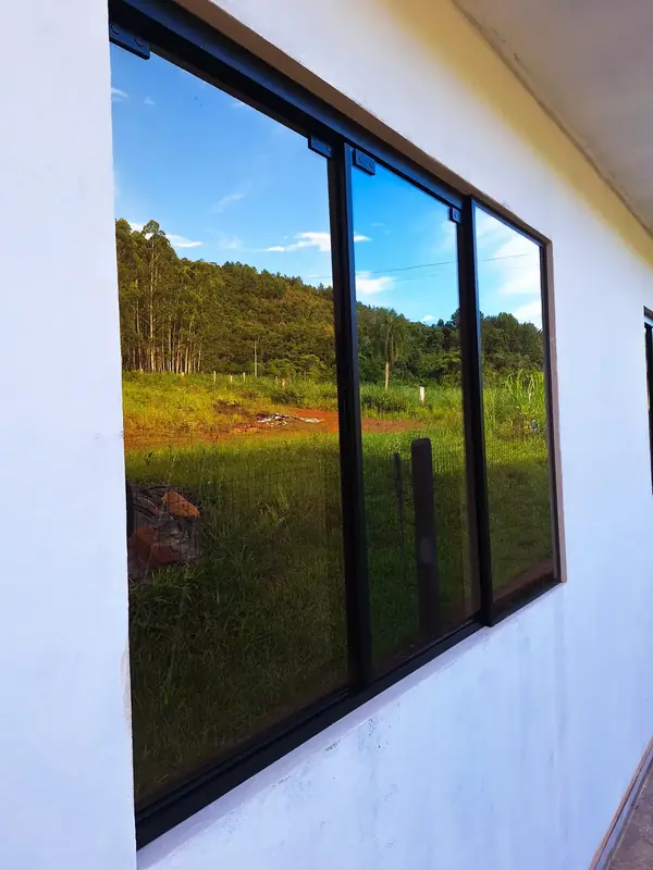 Janela de alumínio preto de três folhas em parede branca, refletindo o campo