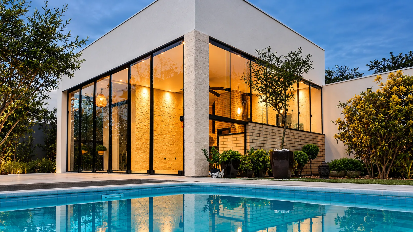 Residência com fachada envidraçada do piso ao teto, iluminada ao anoitecer, vista a partir da piscina